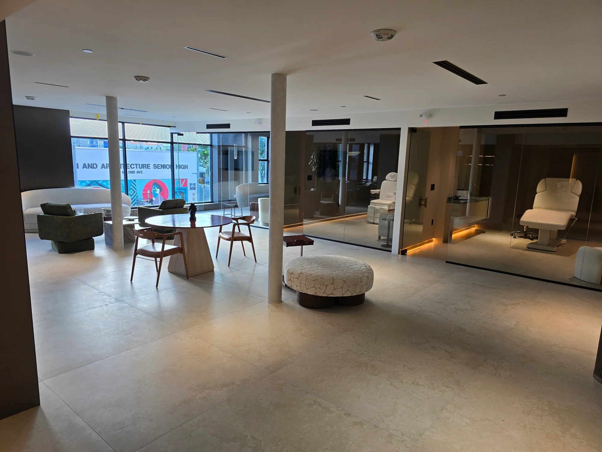 A light-filled lounge with tables, chairs and glass-fronted rooms containing white adjustable chairs.
