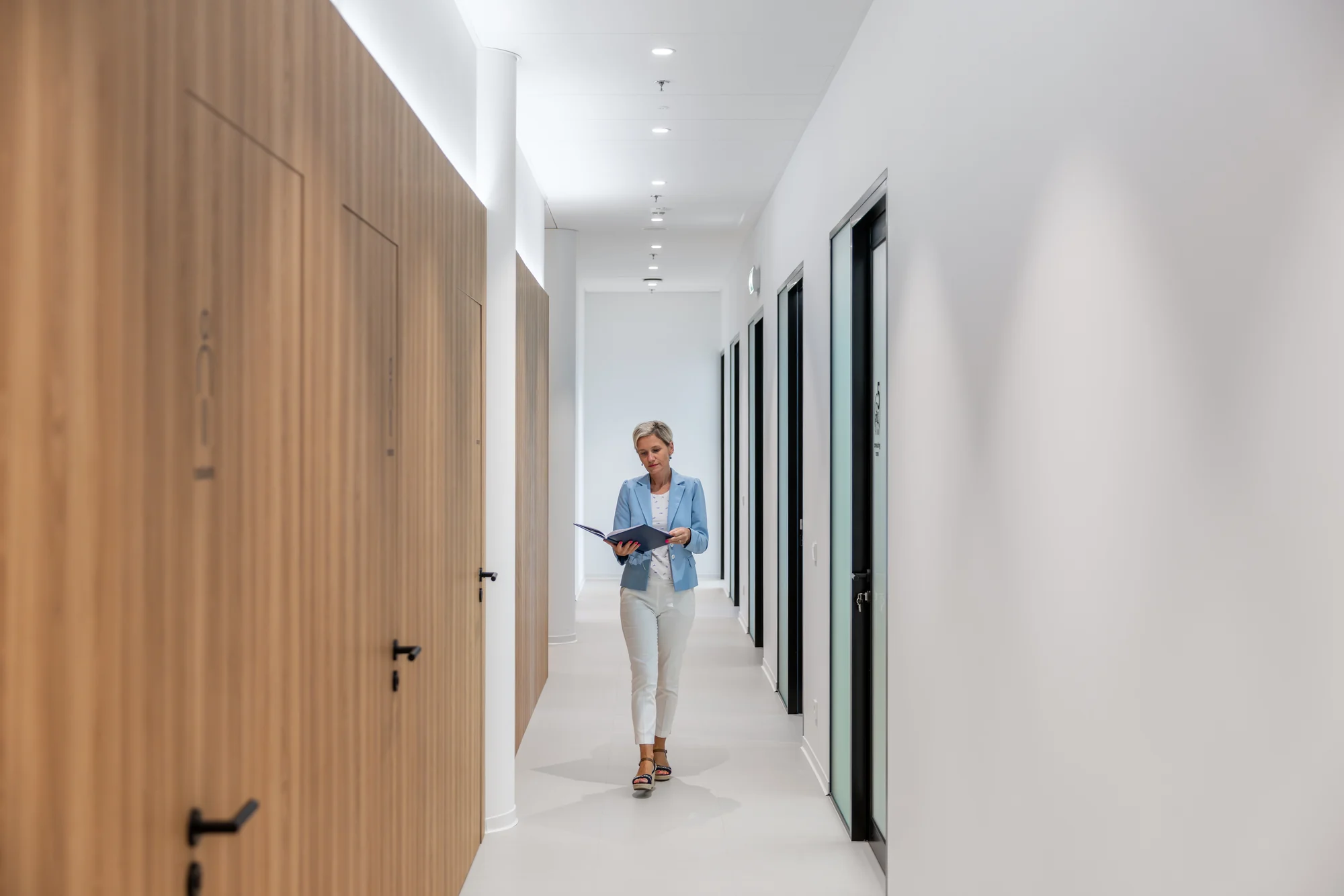 A bright clinic corridor with wood-panelled doors and a woman holding a folder.