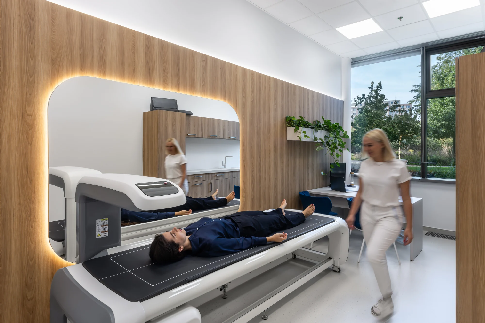 A woman lying on an examination table beside a woman in a white uniform in a bright clinic room.